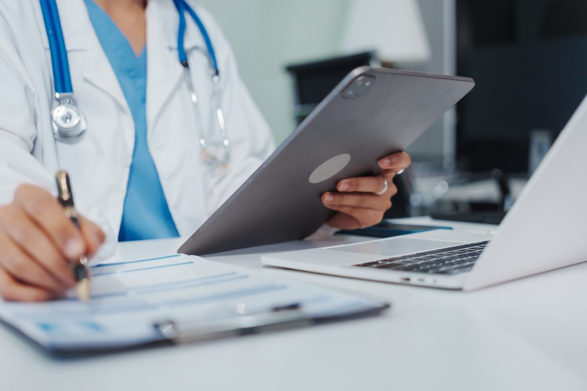 A clinician reviews practice paperwork beside a tablet and laptop.